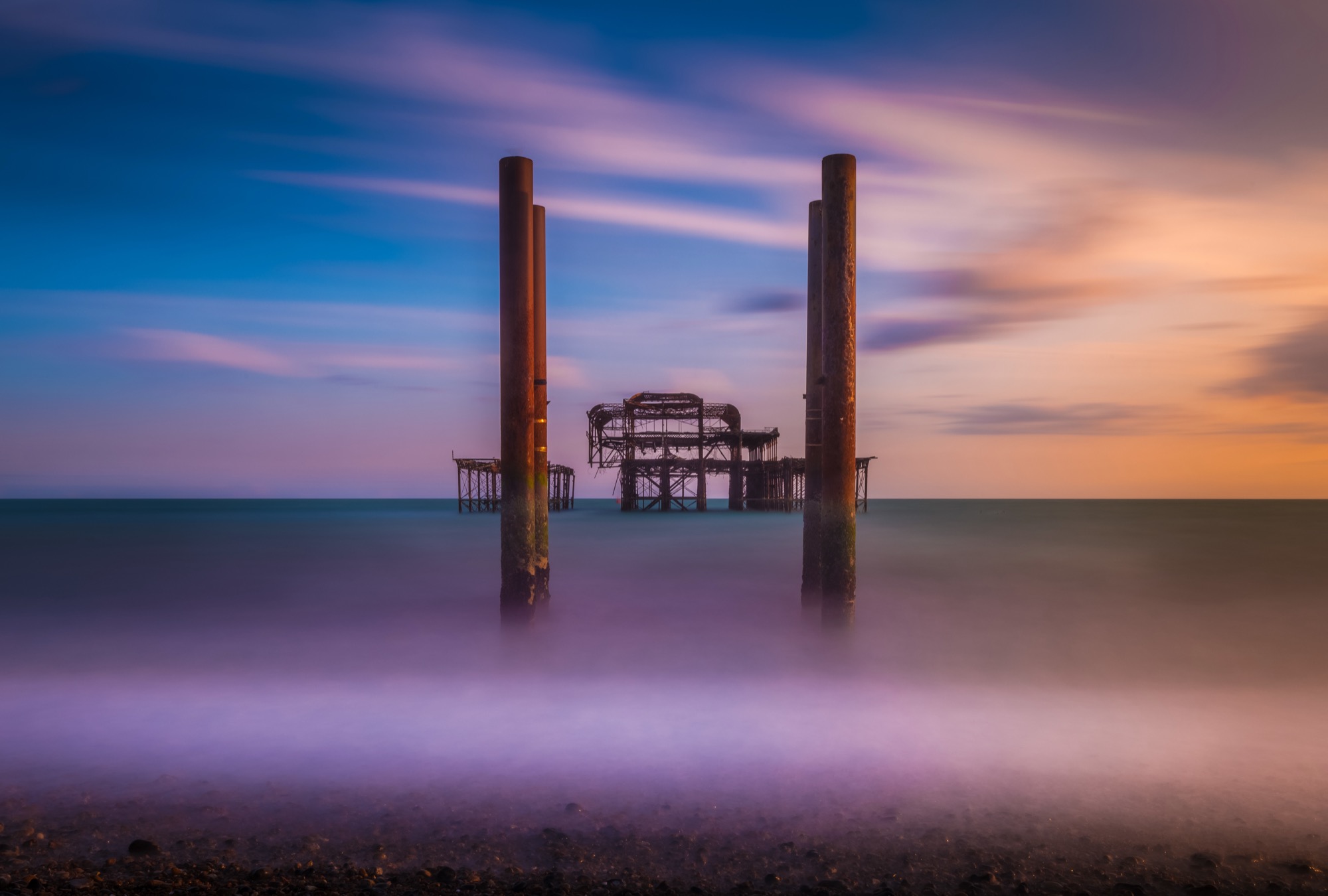 The skeletal iron remains of Brighton's West Pier standing in a calm sea under a pastel sky.