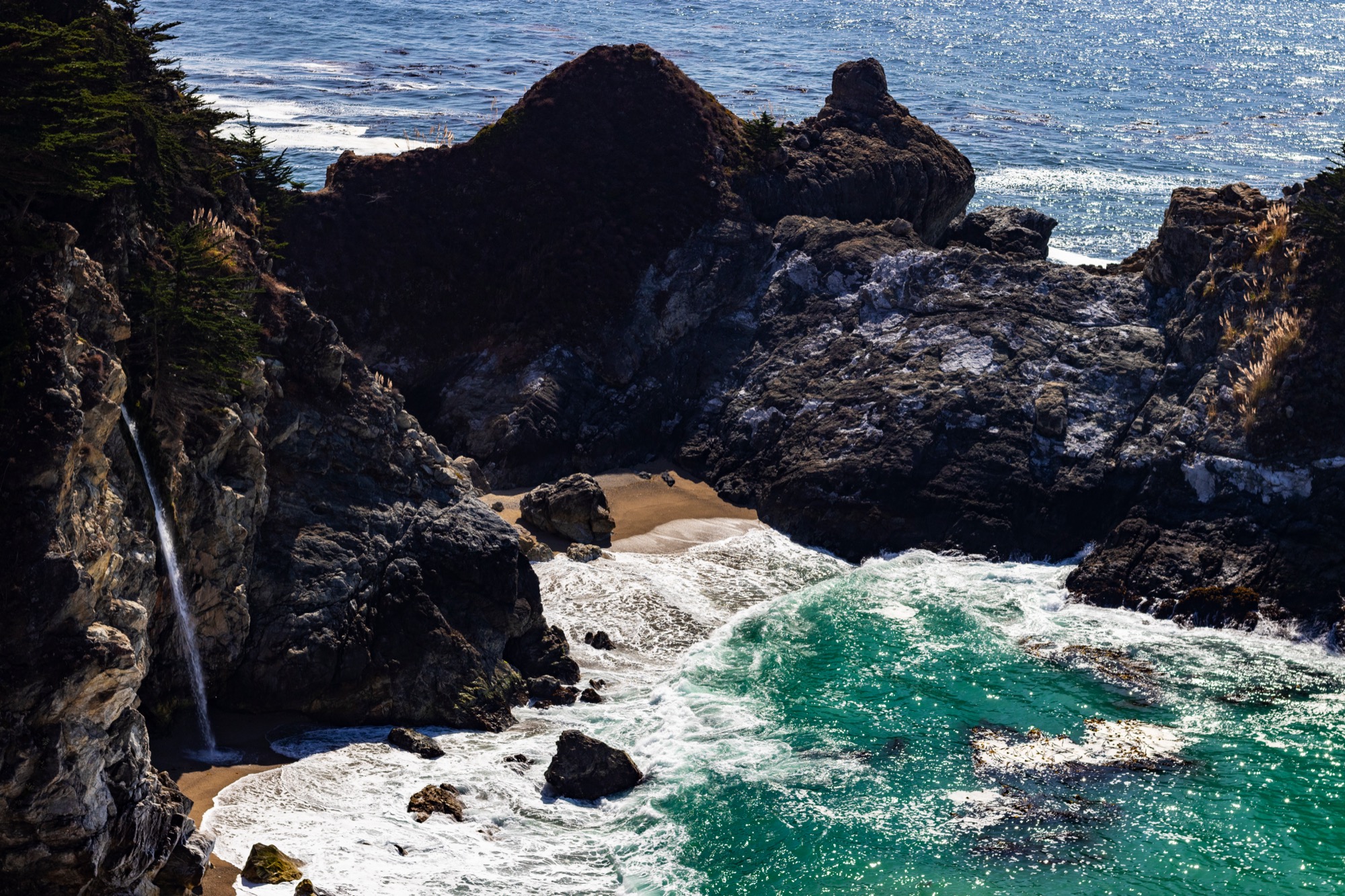 A waterfall falling from a cliff onto a small sandy cove with turquoise sea, on the California coast.