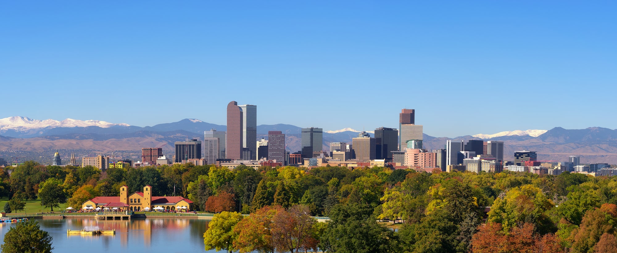 A panoramic view across a city park lake to the Denver skyline, with the snow-covered Rocky Mountains along the horizon behind it.