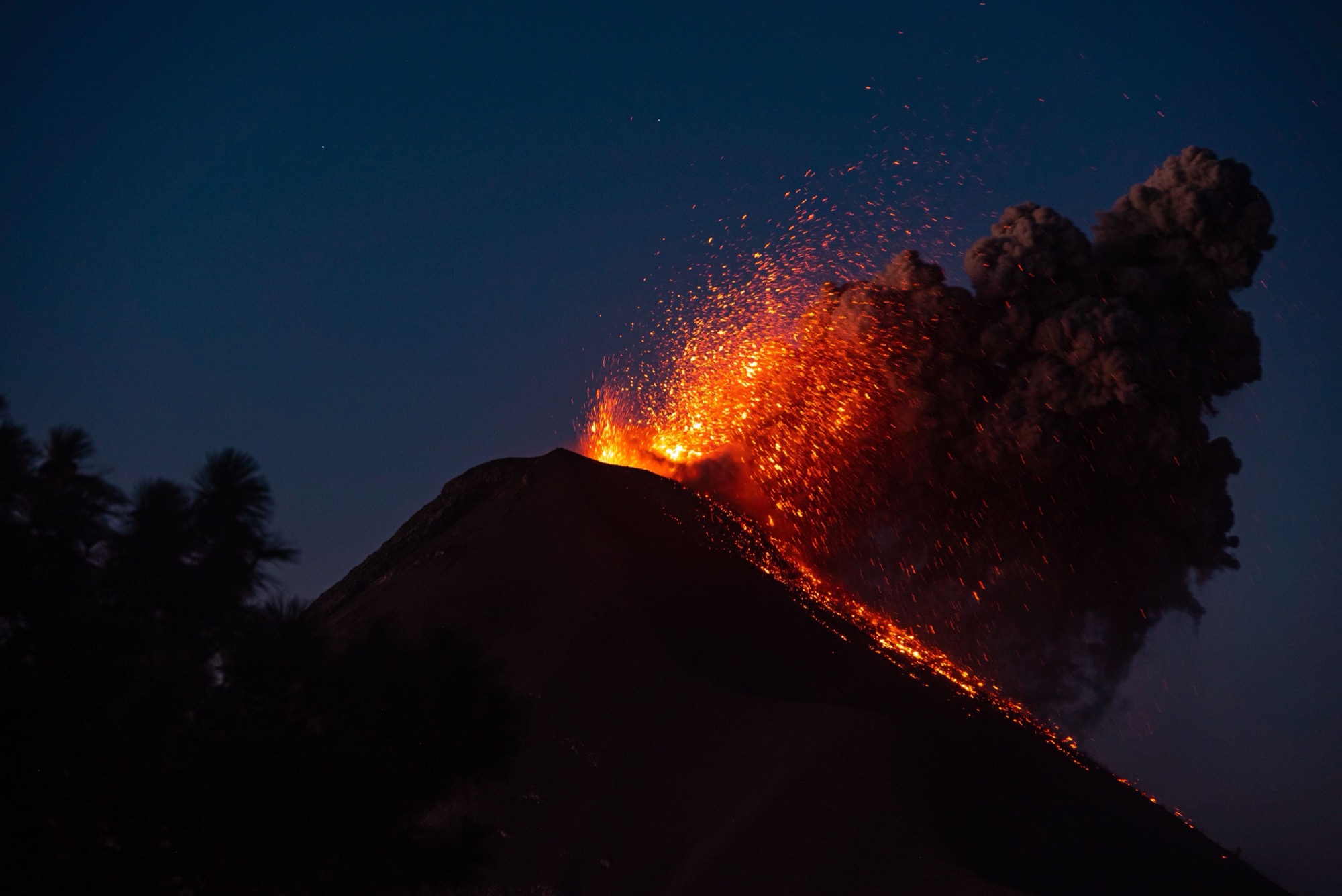 A volcano erupting at night, throwing glowing lava into the air above a dark ash plume.
