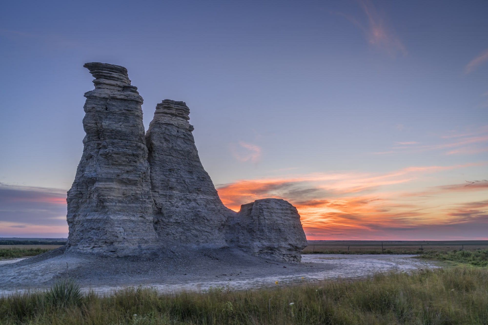 A tall pale limestone pillar rising out of flat prairie grassland, lit by a pink and orange sunrise sky.