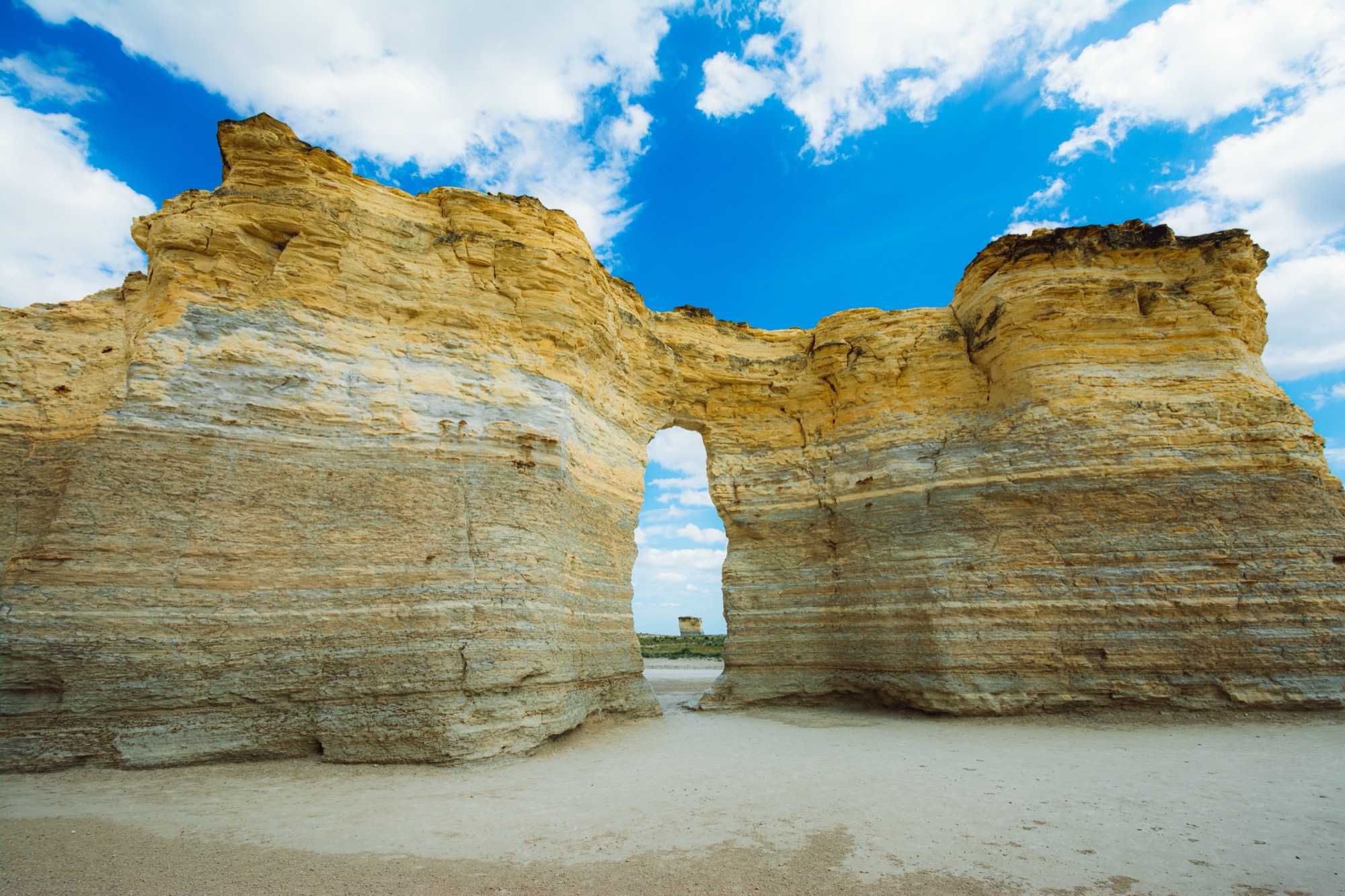 A tall layered wall of pale yellow chalk with an arch through it, standing on flat white ground under a blue sky with clouds.