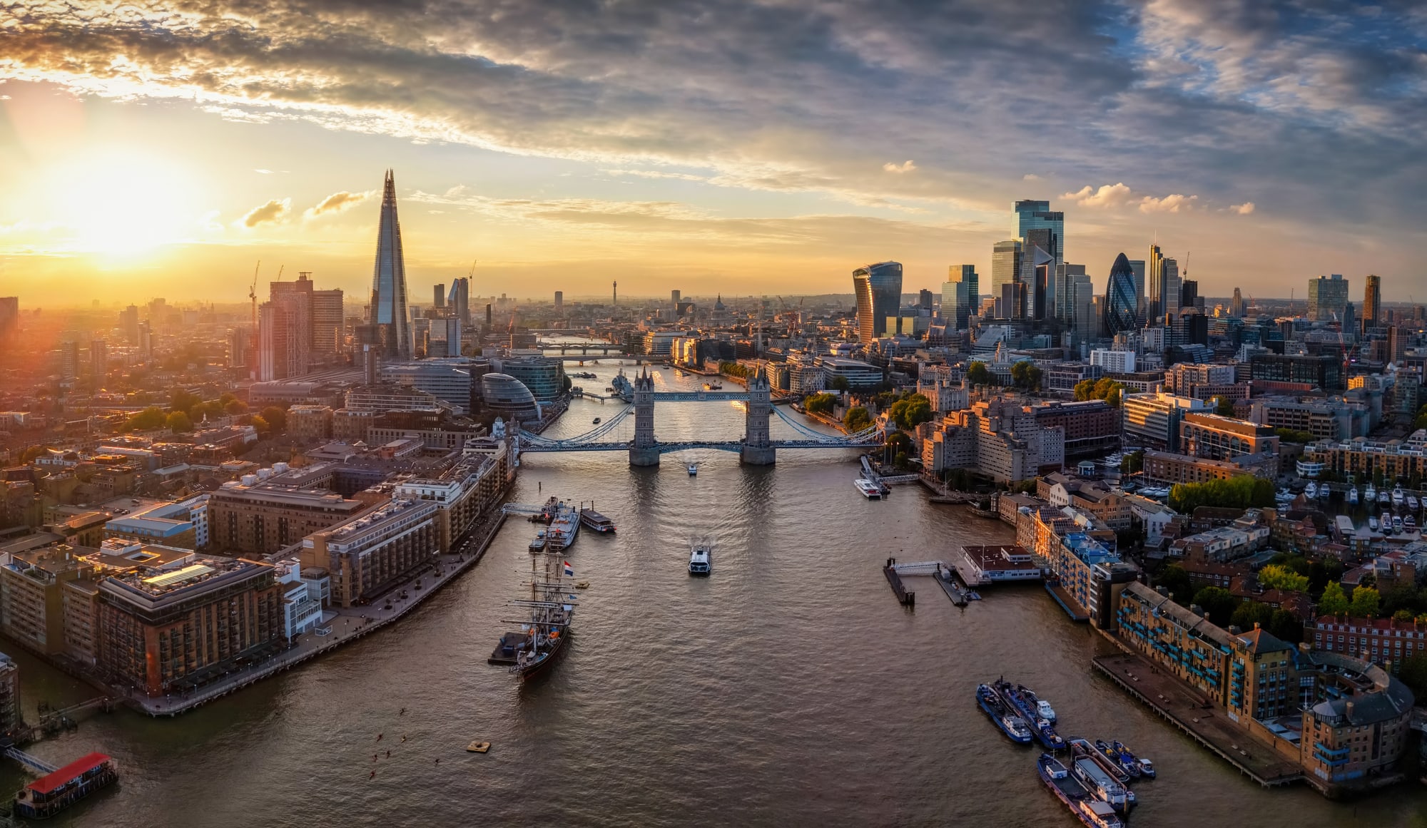 An aerial view along the River Thames at sunset, with Tower Bridge in the middle distance and the towers of the City of London beyond.