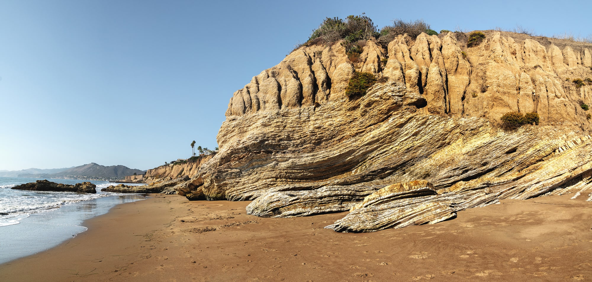A sea cliff of steeply tilted rock layers meeting a sandy beach, with the Pacific Ocean and a headland beyond.