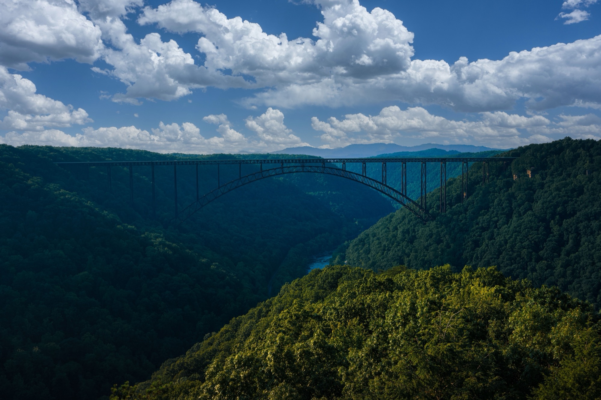 An aerial view of the New River Gorge Bridge, a long steel arch spanning a deep forested gorge, under a partly cloudy blue sky.